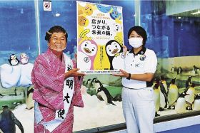 ペンギンが暮らす場所の前で「社会を明るくする運動」のポスターを贈る桂枝曾丸さん（左）＝２６日、和歌山県白浜町のアドベンチャーワールドで