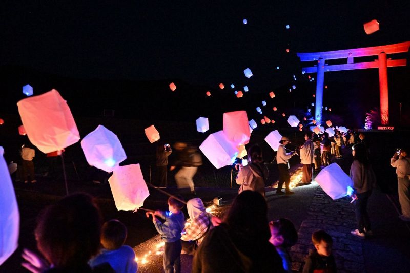 万博カラーにライトアップされた大鳥居と赤と青のランタンを夜空に浮かべる参加者（２３日、和歌山県田辺市本宮町で）