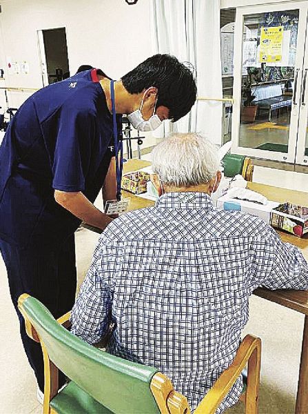 白浜はまゆう病院リハビリテーションセンターでトレーニング問題を解く高齢者に付き添う下村大輔さん（左）＝和歌山県白浜町で
